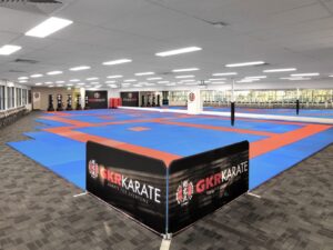 Inside a spacious karate dojo with a large matted area of blue and red. The walls are adorned with banners bearing the GKR Karate logo, and the space is well-lit with rows of fluorescent lights. Along one side of the room, there are several heavy bags for training. The far end has a mirrored wall, enhancing the room's brightness and sense of space. The dojo conveys a professional and welcoming environment for martial arts training, with "Karate for Everyone" as its motto.
