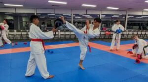 Two karate students are practicing in a dojo with blue and red mats. The student in the foreground, wearing a red belt, holds a kick pad while the other student, wearing a white gi with a red and white belt, executes a high roundhouse kick. In the background, other students can be seen training with pads and gloves, some in the middle of their own exercises. The space looks like a dedicated martial arts training area, with mirrors along the wall that reflect the activity in the room.