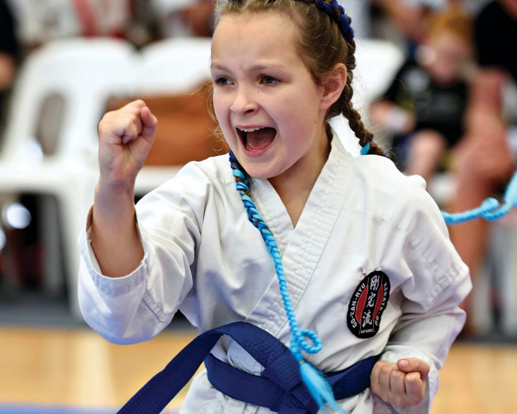 a little girl wearing a blue belt performs a karate move in front of an audience
