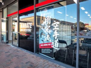 The storefront of a GKR Karate dojo with a large window display. The window features the GKR Karate logo and the words "Karate for Everyone" prominently displayed, alongside an image of a karate practitioner and Japanese characters. There's also advertising text that reads "ZERO JOINING FEE! TAKE ACTION NOW AND SAVE," suggesting a promotional offer. The dojo is situated in a commercial area with other shops and parked cars visible through the glass, reflecting the bustling environment outside. The entrance is framed by black and red design elements, mirroring the branding colors of the dojo.
