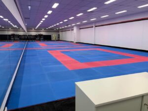 This image shows a wide view of the training area in a martial arts dojo. The floor is covered with a large expanse of interlocking mats in a checkerboard pattern of blue and red. The walls are white with a red stripe running along the top, and the ceiling is fitted with multiple square light panels providing even illumination throughout the space. A portion of a white counter or reception desk is visible in the foreground on the right, giving a perspective of the dojo from the entrance or administrative area.