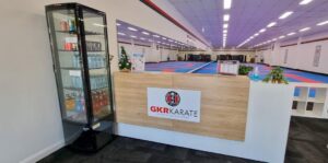 This is the reception area of a GKR Karate dojo, featuring a wooden front desk with the dojo's logo stating "Karate for Everyone". Next to the desk is a refrigerated display case stocked with water bottles and energy drinks. The expansive training area in the background is fitted with blue and red mats, and the space is well-lit with ceiling lights. There's a small Christmas tree and a hand sanitizer bottle on the reception counter, adding a personal touch to the welcoming area.