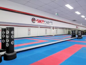 This image shows the interior of a GKR Karate dojo. The space is expansive, with a floor laid out with alternating blue and red mats suitable for martial arts practice. On the left, there are freestanding black punching bags with numbered target areas for precision training. The dojo's walls are clean and bright, adorned with the GKR Karate logo and the phrase "Karate for Everyone", reinforcing the inclusive nature of the dojo. The ceiling is lined with fluorescent lighting, ensuring the space is well-illuminated for training.