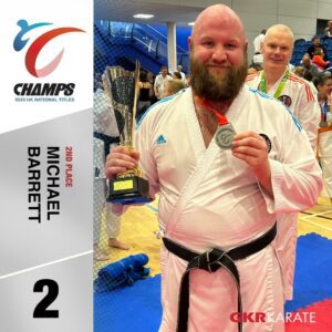 A bearded male karate athlete with a black belt proudly displays his silver medal and holds a black and gold trophy, signifying his second-place finish. He's wearing a white gi with the Adidas logo. In the background, other karate competitors and spectators can be seen in a sports hall with a blue matted floor. The image includes a graphical overlay on the left, with the CHAMPS logo, '2023 UK NATIONAL TITLES', '2ND PLACE', and 'MICHAEL BARRETT' written alongside the number '2'.