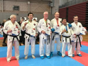 A group of senior male karate practitioners in white gis and black belts stands proudly on a red and blue matted floor, each holding a certificate and wearing a medal. Their cheerful expressions and relaxed postures convey a sense of accomplishment and camaraderie at the karate event.