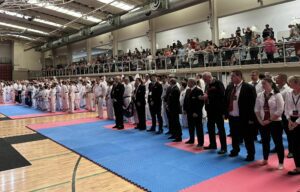 Rows of karate practitioners in white uniforms stand respectfully alongside a group of judges and officials in formal attire and black belts, all facing towards the side of a large indoor sports hall with a blue and red matted floor. Above them, an audience is gathered in the tiered seating area, observing the event with interest
