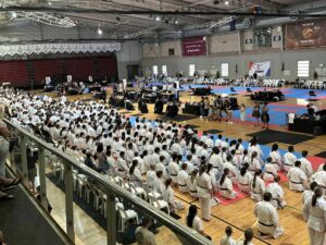 A large indoor sports arena filled with hundreds of karate practitioners seated in white uniforms, as they attentively watch a karate demonstration on the central blue and red mats. Spectators line the balcony above, and banners with branding hang in the background, indicating a significant martial arts event or competition