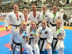 A group of eight karate athletes in white gis, each with a black belt, are kneeling and standing on a blue matted floor, smiling proudly with variously colored medals around their necks. They are in a sports hall with spectators in the stands in the background.