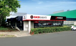 The exterior of a building shared by GKR Karate and POWER Functional Fitness. The karate dojo advertises "Karate for Everyone" and features the GKR Karate logo with Japanese characters. Adjacent is the fitness facility with the POWER branding, emphasizing functional fitness. The building has a modern look with a black and white facade accented with red signage. Reflective windows show the cars parked outside, and the setting includes greenery around the entrance, suggesting an urban location with easy accessibility.