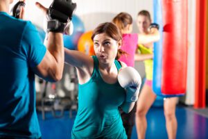 A woman in a teal tank top and white boxing gloves is focused on throwing a punch towards her trainer's padded mitts during a boxing fitness class. In the background, other participants are also engaged in boxing workouts with punching bags. The gym is vibrant, with bright lighting and colourful equipment, suggesting a dynamic and energetic atmosphere.