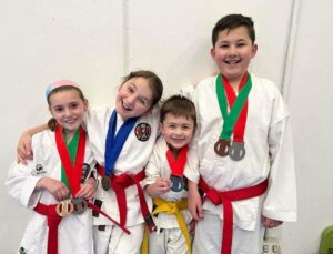 Four young karate students with big smiles, wearing white gi and colored belts, are standing together proudly displaying their medals. The two on the left wear blue and green belts, and their medals are gold and silver, while the two on the right wear red and yellow belts with a silver and two bronze medals. Their cheerful expressions and the medals around their necks suggest they have recently participated in a competition or grading event.