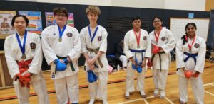 A group of young karate practitioners standing in a gymnasium, equipped with protective hand gear in red and blue. They are wearing white karate uniforms adorned with medals and belts of different colors indicative of their ranks. The students appear to be of various ages and are positioned in a straight line, facing the camera with a display of training posters on the wall behind them.