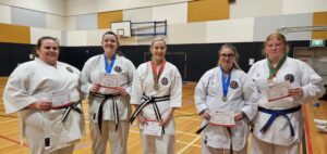 A group of five adult karate students, are lined up in a gymnasium, each holding a 'Certificate of Participation' from GKR Karate. They are dressed in white gis and black belts, with two of the students also wearing medals. The gym has a sporty ambiance with a basketball hoop in the background, and the students are displaying a mix of proud and happy expressions