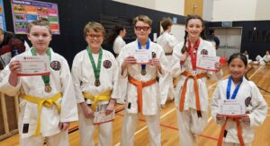 Five young karate students are proudly displaying their 'Certificate of Participation' from a GKR Karate event. They are dressed in white gis with various colored belts, including yellow, orange, and green, and some have medals hanging around their necks. The children are standing in a gymnasium with activity visible in the background, and each student is beaming with pride at their accomplishment