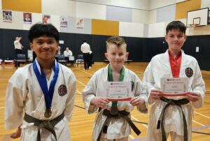 "Three young karate students stand side-by-side, each holding a 'Certificate of Participation' from GKR Karate. The boy on the left with a blue belt and medal smiles at the camera, the boy in the middle with a green belt also holds a bronze medal, and the boy on the right with a red belt poses with confidence. They are in a gymnasium with activity visible in the background