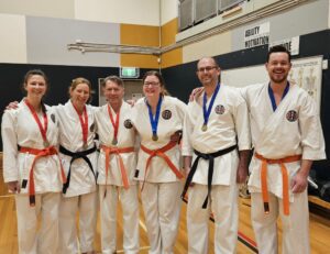 A group of six karate students, three women and three men, smiling and posing together in a dojo. They are wearing white karate gis and colored belts—ranging from orange to black—and are all proudly displaying medals around their necks. A motivational banner with the words 'ABILITY MOTIVATION ATTITUDE' is visible in the background, setting a positive tone in the training environment