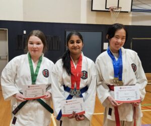 Three young female karate students standing in a gymnasium, each holding a certificate of participation. They are wearing karate gis with their respective belts; green, red, and blue, and medals around their necks. They are smiling at the camera, with a basketball hoop visible in the background.