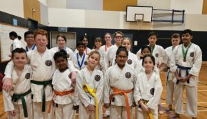 A group of young karate students in a gymnasium, proudly wearing their uniforms and various colored belts, with some displaying medals. They are standing in several rows, smiling for the photo, with a scoreboard and a basketball hoop visible in the background.