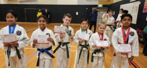A group of six young karate students stands side by side on a gymnasium floor, displaying their participation certificates with a sense of achievement. They are attired in traditional white karate uniforms, called gis, and are adorned with different colored belts, indicating their ranks. A few of them also wear medals, signifying their accomplishments in the competition. Behind them, the gym is bustling with other competitors and observers, creating a backdrop of activity and enthusiasm for martial arts