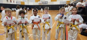 A group of six young karate students, with vibrant smiles, poses for a photo in a gymnasium. They are dressed in white karate gis with colored belts ranging from yellow to orange, signifying their progression in the martial art. Each child proudly holds a certificate of participation, and some are adorned with medals around their necks. The gym is lively with other karate students and spectators in the background, emphasizing the community and supportive atmosphere of the event