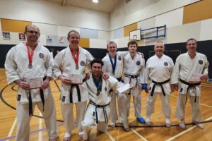 Seven karate students pose with joy after receiving their participation certificates. They are all dressed in white gis with black belts, and several have medals. Each student shows a different expression of happiness and pride. The dojo's walls are adorned with posters, and the gym setting includes a basketball hoop in the background, highlighting the community and sportsmanship spirit of the event