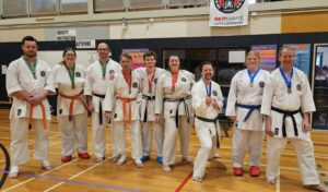 A diverse group of nine adult karate students stands in a line in the gymnasium, some with medals around their necks and certificates in hand. They are wearing white gis with belts of various colors including orange, green, brown, and black. Above them hangs a banner reading 'GKR KARATE NORTH CANTERBURY', and motivational posters adorn the wall in the background. Their expressions range from proud smiles to contented grins, reflecting a sense of achievement