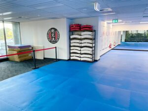 The interior of a dojo with a spacious blue practice mat. To the right, there's a neatly organized storage area with shelves holding folded white gi uniforms and red practice pads. Japanese kanji symbols are displayed on a circular emblem on the wall, adding a cultural touch. The dojo is well-lit with natural light from the windows along the far wall