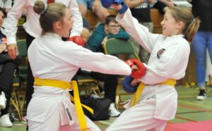 Two young girls in white karate gis and colored belts are sparring. The girl on the left with a yellow belt has thrown a high kick, while the girl on the right with a yellow belt blocks with her forearm. Both wear red protective gloves and are focused on their match, showing great spirit and concentration. In the background, spectators and fellow competitors watch the action