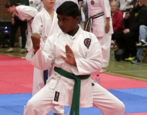 A young boy in a karate stance wears a white gi and a green belt, focused intently as he performs a kata. His fist is clenched near his hip while his other hand is extended in a knife-hand block. He is on a blue and red matted floor with other karatekas practicing in the background and spectators observing the event