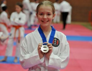 A young girl with a beaming smile, wearing a white karate gi and a blue belt, proudly shows off a silver medal. The medal is held up in front of her with both hands, drawing attention to her achievement. She stands in a dojo with a blurred background where other karate students can be seen on a red and blue matted floor