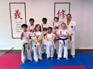 A joyful group of young karate students, along with their instructor, are proudly displaying their achievement certificates. They're standing on a red and blue matted floor in front of white walls adorned with large Japanese kanji calligraphy. The banners read ‘Pursue the highest standard of personal character’ and ‘Cultivate,’ reinforcing the dojo's values
