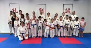 A diverse group of smiling karate students of various ages and belt colors, along with their instructors, are posing for a group photo on a blue and red matted floor in the dojo. Behind them are walls decorated with Japanese kanji characters on white banners