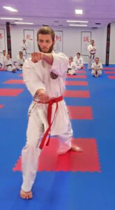 A focused male karate student with a red belt is in a forward stance throwing a punch towards the camera. He is in a dojo with a blue and red matted floor, and other students are seated in the background observing, under Japanese kanji character wall banners