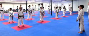 A diverse group of karate students in white uniforms and different colored belts standing in rows, ready to begin practice on a blue and red matted floor in a large dojo. Traditional Japanese calligraphy banners decorate the walls, creating an authentic martial arts atmosphere