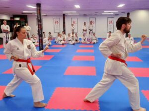 Two karate students, a woman and a man, in white gis with red belts, are practicing kata with focused expressions in the center of a dojo. In the background, other students seated on a blue and red matted floor watch attentively, surrounded by traditional Japanese kanji characters on the wall banners