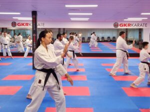 A focused karate class in session with multiple students, including a woman in the foreground, performing synchronized kata movements on a blue and red matted floor in a well-lit dojo, with the GKR Karate logo visible in the background