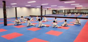 Karate students of various ages and ranks are sitting and stretching on a blue and red matted floor in a spacious dojo. GKR Karate banners line the practice area, adding to the focused yet relaxed atmosphere of the class